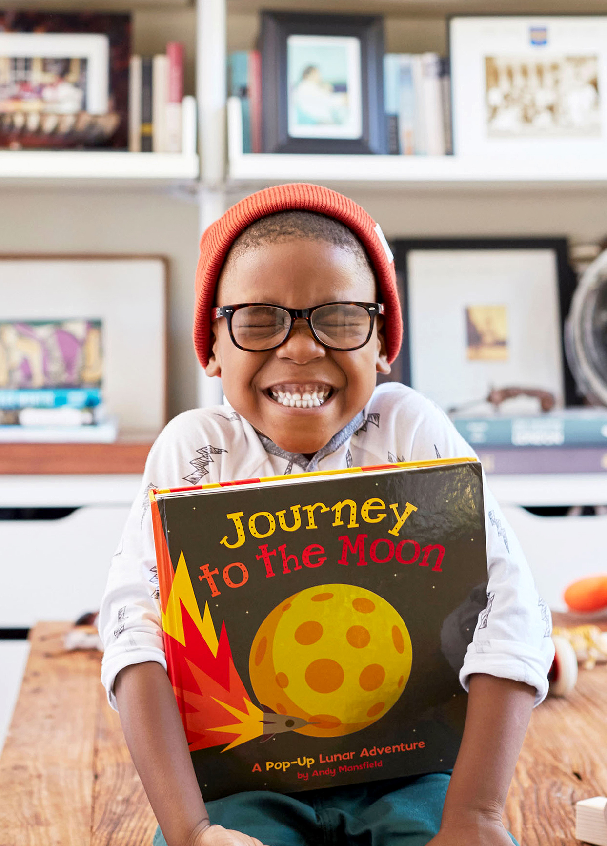 smiling preschool boy sitting on coffee table with glasses, hat, and book