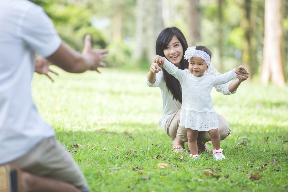 Toddler Learning To Walk With Parents Outside On Grass